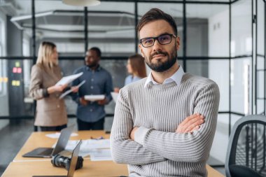 Adam kollarını kavuşturmuş duruyor. Profesyonel işçiler modern ofiste birlikte çalışıyorlar..