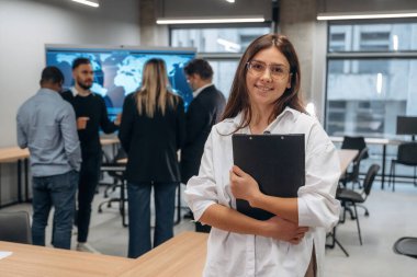 Kadın kameraya bakıyor. Bir grup ofis çalışanı içeride birlikte..