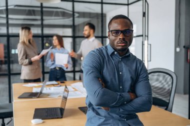 Siyah adam ayakta duruyor ve poz veriyor. Profesyonel işçiler modern ofiste birlikte çalışıyorlar..