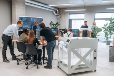 Duvarında kod olan büyük ekran. Ofiste 3D yazıcının yanında insanlar var..