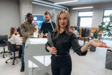 Genç bayan meslektaşlarının önünde duruyor. Ofiste 3D yazıcının yanında insanlar var..