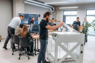 Duvarında kod olan büyük ekran. Ofiste 3D yazıcının yanında insanlar var..