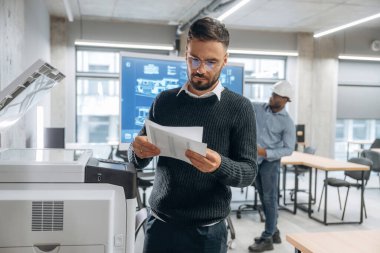 İki ofis çalışanı ayakta. Yazıcının yanında belgeleri olan bir adam var..