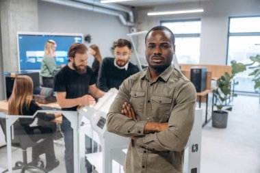 Afrikalı Amerikalı bir adam meslektaşlarının önünde duruyor. Ofiste 3D yazıcının yanında insanlar var..