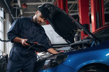 Elinde siyah dijital tablet var. Araba tamirhanesinde çalışan bir tamirci..