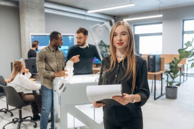 Kadın meslektaşlarının önünde duruyor. Ofiste 3D yazıcının yanında insanlar var..