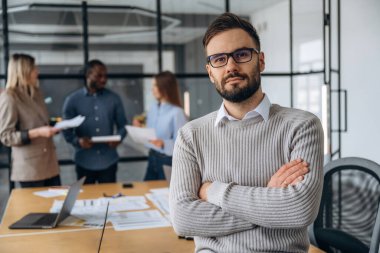 Adam kollarını kavuşturmuş duruyor. Profesyonel işçiler modern ofiste birlikte çalışıyorlar..