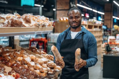 Çeşit çeşit gösteriliyor. Süpermarket işçisi ekmek ürünlerinin yanında duruyor.