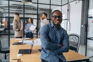 Siyah adam ayakta duruyor ve poz veriyor. Profesyonel işçiler modern ofiste birlikte çalışıyorlar..