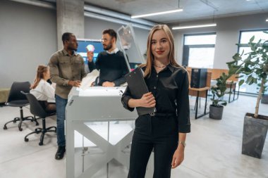 Elinde not defteri var, kadın ayakta. Ofiste 3D yazıcının yanında insanlar var..