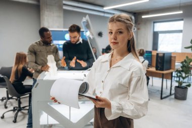 Elinde not defteri var, kadın ayakta. Ofiste 3D yazıcının yanında insanlar var..