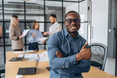 Siyah adam ayakta duruyor ve poz veriyor. Profesyonel işçiler modern ofiste birlikte çalışıyorlar..