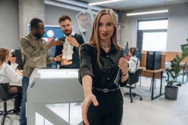 Genç bayan meslektaşlarının önünde duruyor. Ofiste 3D yazıcının yanında insanlar var..