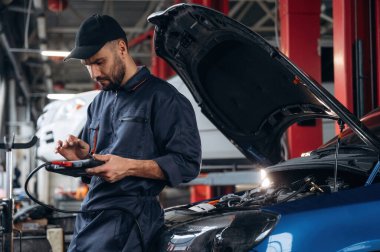 Elinde siyah dijital tablet var. Araba tamirhanesinde çalışan bir tamirci..