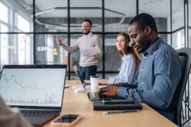 Odaklanmış, dizüstü bilgisayarın yanında oturuyor. Profesyonel işçiler modern ofiste birlikte çalışıyorlar..