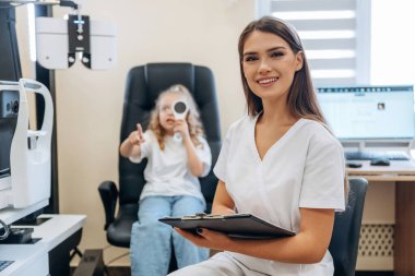 Oturuyor ve not defteri tutuyor. Göz doktoru kliniğinde doktorla küçük bir kız..