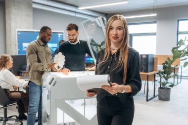 Kadın meslektaşlarının önünde duruyor. Ofiste 3D yazıcının yanında insanlar var..