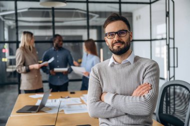 Adam kollarını kavuşturmuş duruyor. Profesyonel işçiler modern ofiste birlikte çalışıyorlar..