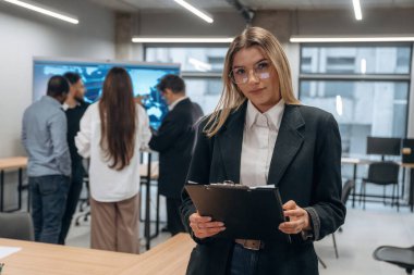 Gözlüklü kadın elinde not defteriyle ayakta duruyor. Bir grup ofis çalışanı içeride birlikte..