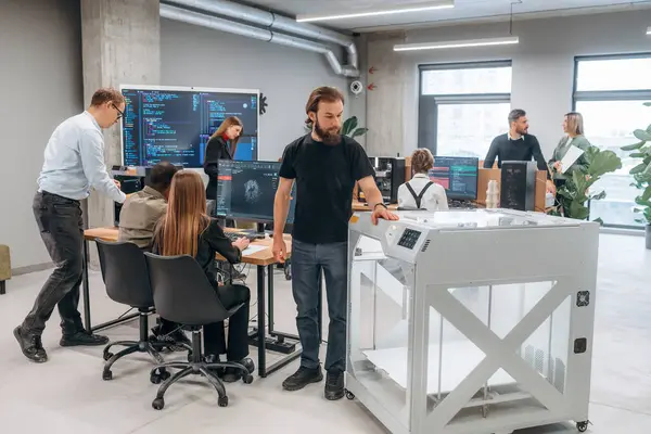 Duvarında kod olan büyük ekran. Ofiste 3D yazıcının yanında insanlar var..