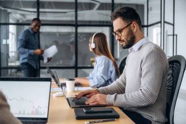 Odaklanmış, dizüstü bilgisayarın yanında oturuyor. Profesyonel işçiler modern ofiste birlikte çalışıyorlar..