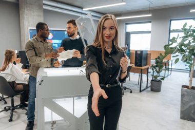 Genç bayan meslektaşlarının önünde duruyor. Ofiste 3D yazıcının yanında insanlar var..