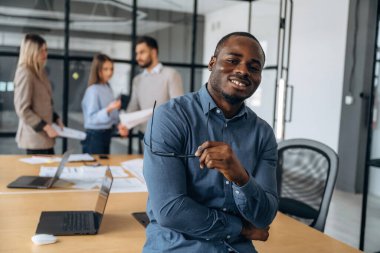 Siyah adam ayakta duruyor ve poz veriyor. Profesyonel işçiler modern ofiste birlikte çalışıyorlar..