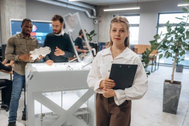 Elinde not defteri var, kadın ayakta. Ofiste 3D yazıcının yanında insanlar var..