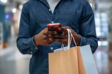 Akıllı telefondan interneti kaydırmak. Süpermarketteki alışveriş çantalı adamın yakın görüntüsü..