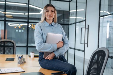 Poz vermek, masada oturmak, dizüstü bilgisayarı tutmak. Kadınlar modern ofiste, resmi kıyafetler giyiyorlar..