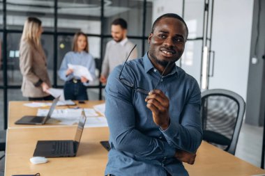Siyah adam ayakta duruyor ve poz veriyor. Profesyonel işçiler modern ofiste birlikte çalışıyorlar..