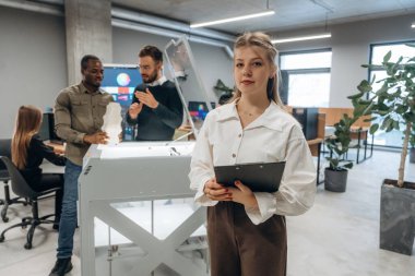 Elinde not defteri var, kadın ayakta. Ofiste 3D yazıcının yanında insanlar var..