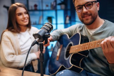 Akustik gitarla. Radyodaki erkek müzisyen kadın sunucuyla podcast yapıyor..