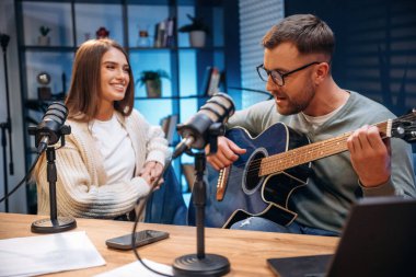 Akustik gitarla. Radyodaki erkek müzisyen kadın sunucuyla podcast yapıyor..
