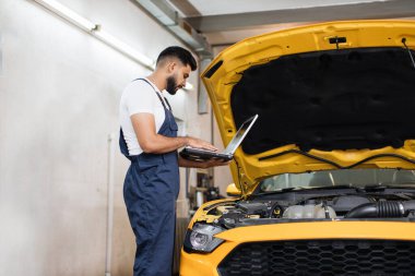 Bearded male mechanic using laptop, recording automobile engine checks collect detailed information during his work on car workshop. Service maintenance during engine repair. clipart