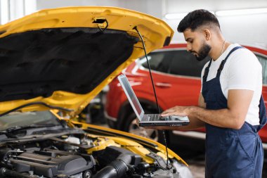 Sakallı erkek tamirci dizüstü bilgisayar kullanıyor, otomobil motor kontrollerini kaydediyor araba atölyesindeki işi sırasında detaylı bilgi topluyor. Motor onarımı sırasında servis bakımı.