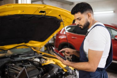 Ciddi odaklı erkek araba teknisyeni motor tamir sorunu, sistem kontrolü sırasında, araba garajında bakım ve onarım için tablet bilgisayar kullanılıyor.