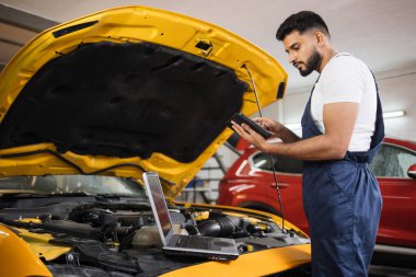 Ciddi odaklı erkek araba teknisyeni motor tamir sorunu, sistem kontrolü sırasında, araba garajında bakım ve onarım için tablet bilgisayar kullanılıyor.