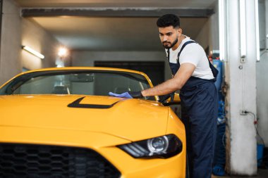 Araba yıkayan bir adamın tam boy portresi koruyucu kıyafet üniforması giyiyor, modern sarı lüks arabayı mikrofiber bezle temizliyor. Araba temizleme veya araç konsepti.