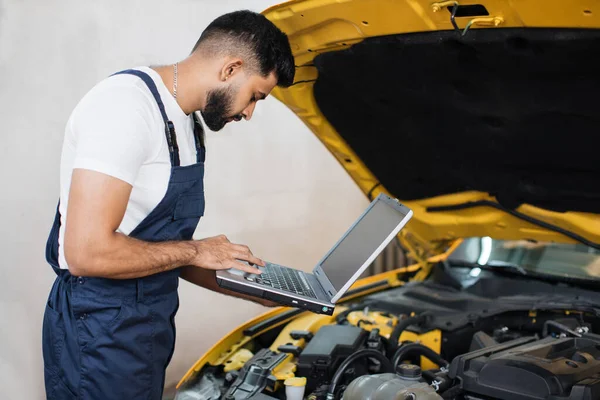 Bearded male mechanic using laptop, recording automobile engine checks collect detailed information during his work on car workshop. Service maintenance during engine repair.