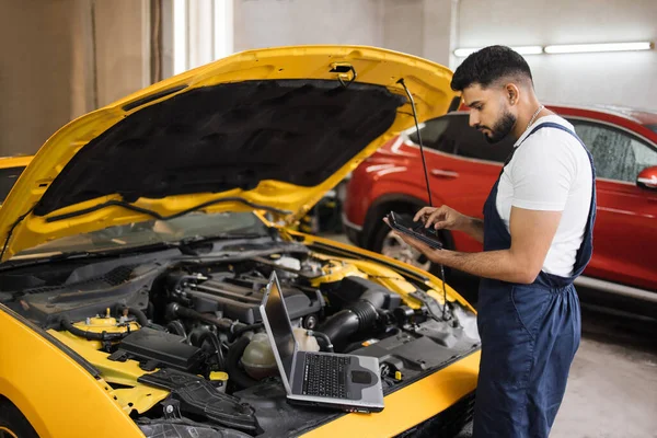 Araba servis istasyonu. Genç sakallı çekici tamirci, motor arızalarını kontrol etmek için tablet ve dizüstü bilgisayarla çalışırken arabanın kaputunun altına bakıyor..