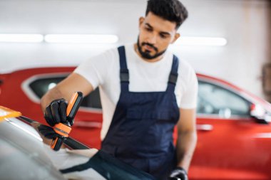 Mavi tulumlu, koruyucu eldivenli, profesyonel bir araba yıkama servisine odaklan. Lüks sarı arabanın ön camına yağmur önleyici sür..