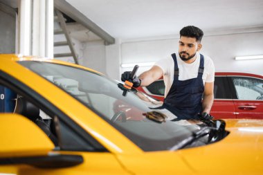Mavi tulumlu ve koruyucu eldivenli erkek sakallı işçi profesyonel bir araba yıkama servisinde, lüks sarı arabanın ön camına yağmur önleyici sürüyor..