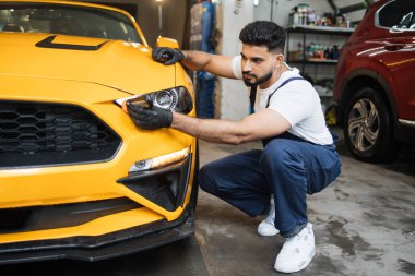 Mavi tulumlu ve koruyucu eldivenli erkek sakallı işçi profesyonel bir araba yıkama servisinde lüks sarı bir arabanın farına yağmur önleyici sürmüş..