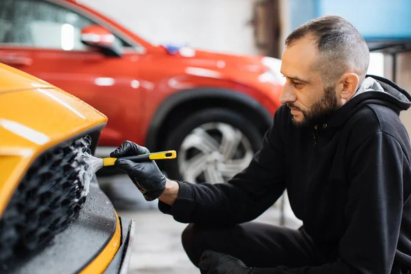 stock image Car detailing wash. Manual cleaning with soap at car wash. Portrait of bearded male worker in rubber glove washing car radiator grille of luxury yellow car with special brush and cleansing foam.