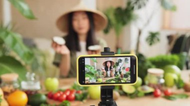 Akıllı telefona odaklan. Geleneksel konik şapkalı Asyalı güzellik blogcusu kahvaltıda meyve püresi yapmak için hindistan cevizi kullanıyor ve çevrimiçi kanal için video kaydediyor..
