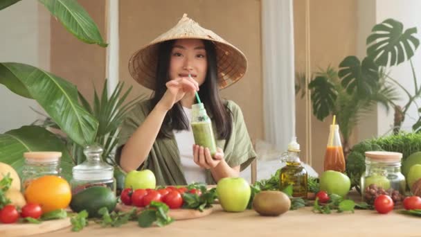 美丽的亚洲女人 戴着传统的锥形礼帽 混合水果和蔬菜配料 在时尚的热带厨房里 坐在餐桌旁做着光滑的食物 年轻女子饮用新鲜绿滋补品 — 图库视频影像