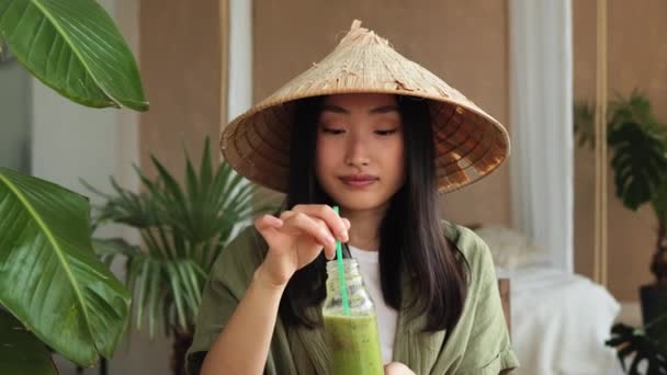年轻的亚洲女人戴着传统的锥形礼帽 用稻草从瓶中喝着健康的滋补品冰沙 而坐在室内的时尚淡淡的热带工作室的绿色新鲜配料桌面上 — 图库视频影像