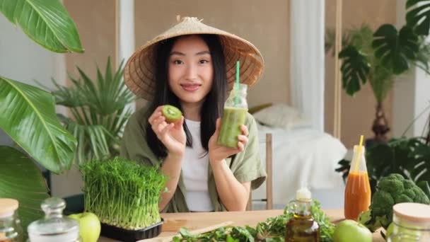 若いですアジアの女性で円錐形の帽子保持で彼女の手ガラス瓶で健康的な強壮スムージーとキウイながら テーブルの上に座っている緑の新鮮な食材室内でスタイリッシュな光トロピカルスタジオ — ストック動画
