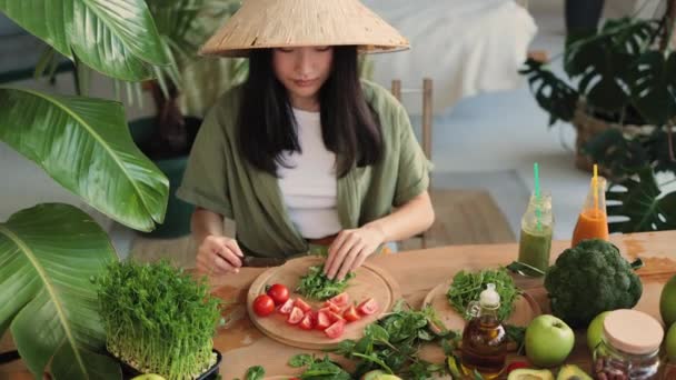 伝統的な帽子の美しいアジアの若い女性の高い角度のビューは 多くの新鮮な果物や野菜と明るいエキゾチックなスタジオでテーブルに座って Arugulaの葉を切っている調理の準備をしています — ストック動画